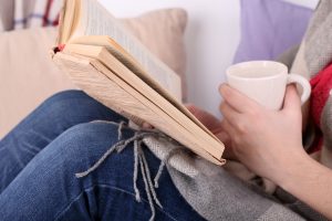 Woman sitting on sofa, reading book and drink coffee or tea, c