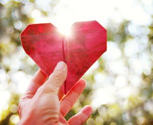 a hand holding an origami paper heart up to the sun during sunse